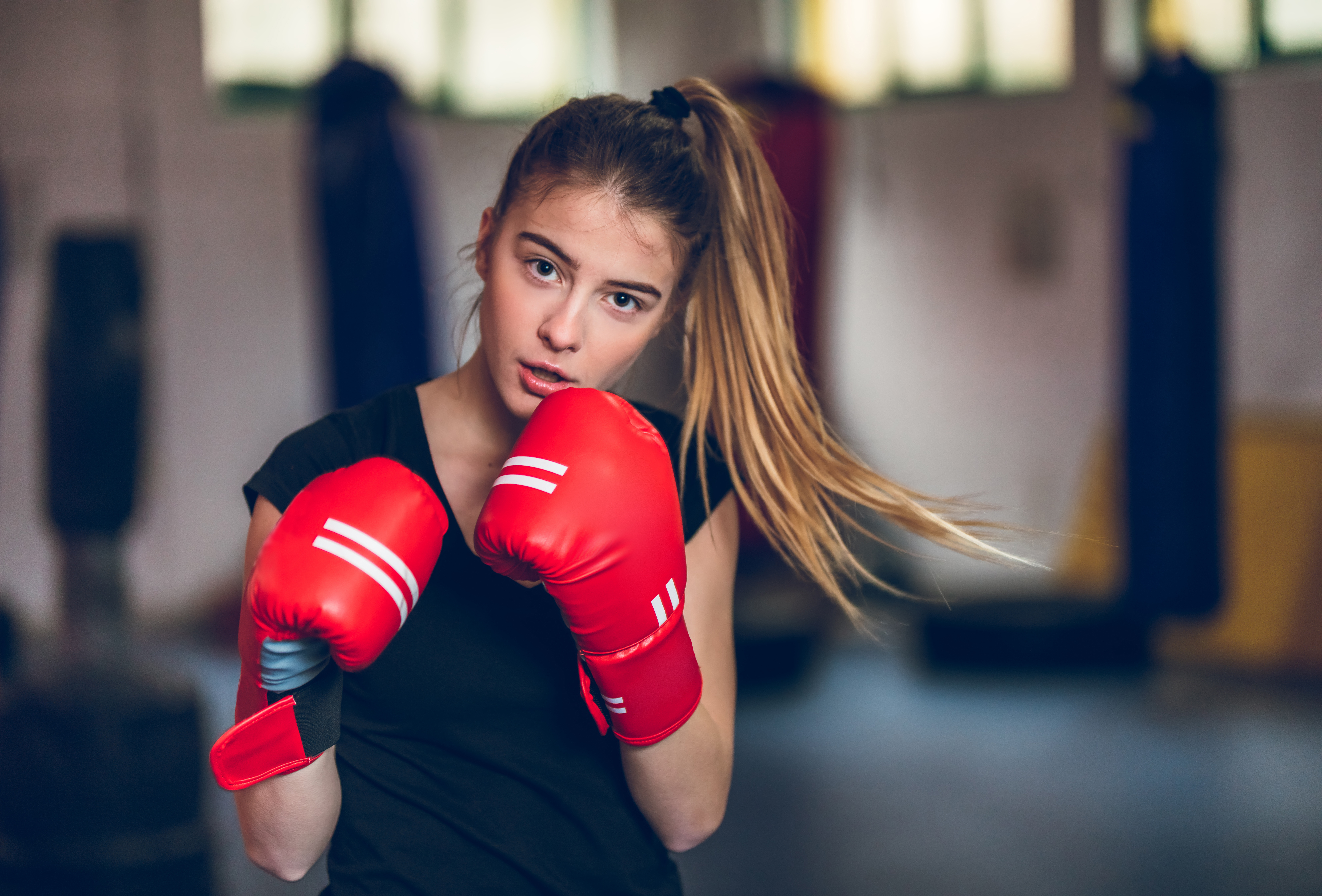A teenage girl with a ponytail and red boxing gloves on. She is holding her hands up to her face.